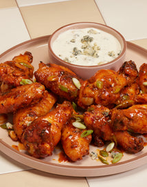A plate of saucy buffalo chicken wings garnished with chopped green onions is served with a bowl of creamy blue cheese dipping sauce on a checkered tan and white surface.