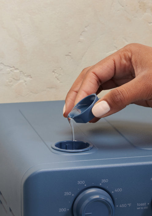 A hand pours liquid from a small blue cup into the top opening of a blue kitchen appliance with a temperature dial on the front.