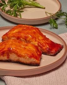 Two pieces of glazed salmon with a shiny, orange-red sauce are served on a light pink plate. Fresh green herbs and another plate are in the background on a light surface.