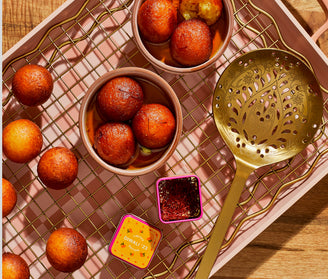 A tray with bowls of gulab jamun, a South Asian dessert, surrounded by more gulab jamun, a gold slotted spoon, and a small container of saffron, all set on a wire rack over a pink tray.