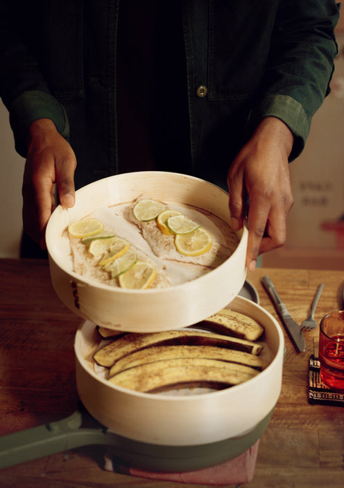 A person holds a bamboo steamer tray with fish fillets topped with lemon slices above a pan containing whole ripe bananas on a wooden table.