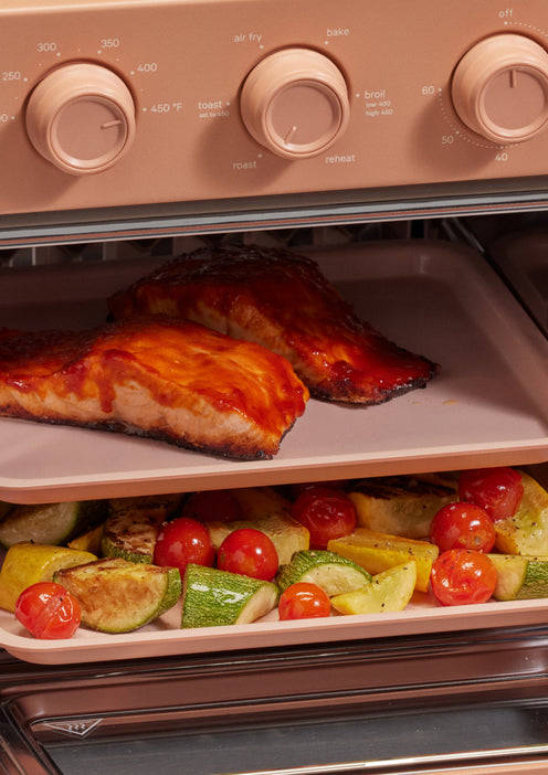 Two trays in an oven: the top tray holds two pieces of glazed salmon, and the bottom tray has roasted vegetables including zucchini, yellow squash, and cherry tomatoes.