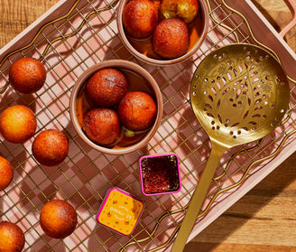 Golden-brown gulab jamuns in small bowls and on a cooling rack, with a decorative slotted spoon and a small container of saffron on a pink tray over a wooden surface.