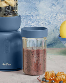 A blue blender with a glass jar of smoothie beside it. The jar contains a purple-blue smoothie. Nearby are fresh blueberries, banana, and a honeycomb on a plate. A bowl with a banana is in the background.