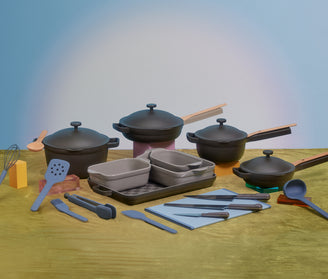 A variety of kitchen cookware and utensils, including pots with lids, baking dishes, trays, spatulas, tongs, a whisk, and a ladle, are arranged on a green surface against a blue background.