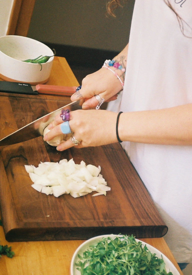 A person wearing rings and bracelets is chopping an onion on a wooden cutting board. Fresh herbs and vegetables are visible on the table, along with bowls and a knife.