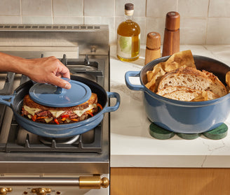A person places a blue lid on a pot containing a sandwich with melted cheese and vegetables on a stove. Next to it, a blue pot on a counter holds sliced bread and parchment paper.