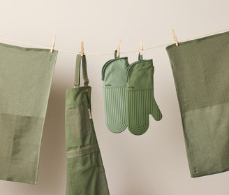 Green kitchen apron, oven mitts, and tea towels hang from a clothesline with wooden clothespins against a neutral background. The items are neatly arranged and color-coordinated in various shades of green.