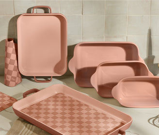 A set of pink ceramic baking dishes and a matching checkered pitcher are arranged on a marble countertop against a tiled kitchen wall. The dishes vary in size, with one featuring a pink checkered pattern.