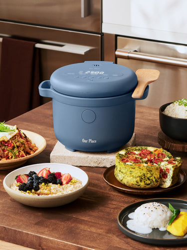A blue electric multicooker sits on a kitchen counter surrounded by various prepared dishes, including rice, vegetables, and a frittata.