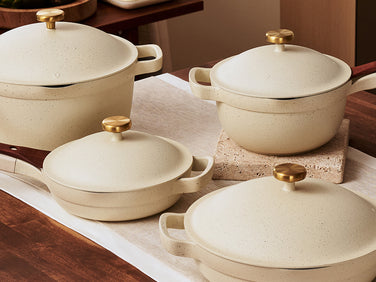 Four beige pots and pans with gold knobs and wooden handles are arranged on a wooden kitchen counter with a table runner underneath.