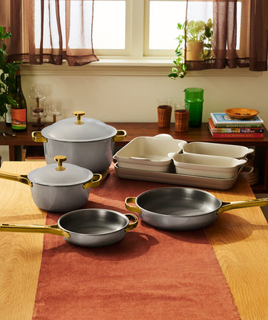 A set of grey cookware with gold handles, including a lidded Dutch oven, a lidded saucepan, two skillets, and a stack of three rectangular baking dishes on a wooden table.