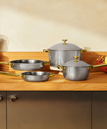 A set of gray cookware with gold-toned handles and lids arranged on a wooden table.