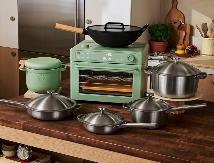 A kitchen countertop with stainless steel cookware, a green multi-cooker, a green toaster oven, a wok, and vegetables on a cutting board in the background.