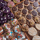 Assorted cookies on cooling racks, including chocolate, frosted, sprinkled, and colorful varieties arranged in neat rows.