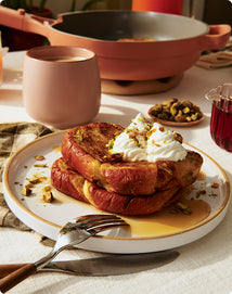 Stack of pancakes with whipped cream and nuts on a plate, served with a cup of coffee and a pan in the background.