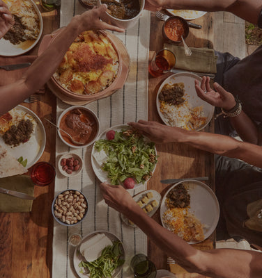 A table set for a meal with dishes of salad, nuts, olives, bread, and various foods. Two people’s hands are reaching for food. Plate and glassware are visible on a woven placemat.