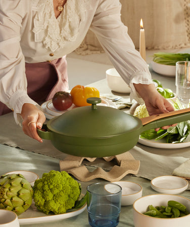 A person places a green, lidded pan with a wooden handle on a trivet at a dining table set with plates, vegetables, cups, and candles in warm natural light.