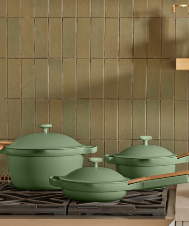 Three green pots and pans with wooden handles sit on a black stove in front of a wall with vertical olive-green tiles in a modern kitchen.
