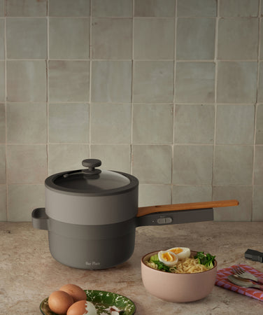 A modern gray and black pot with a wooden handle sits on a kitchen counter. Next to it is a bowl of ramen with vegetables and a boiled egg, a fork, and a yellow vase in the background.