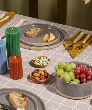 A neatly set dining table with gray plates holding slices of bread and pear, yellow napkins, assorted grapes, nuts, and snacks in bowls, three candles in blue, green, and orange, and a vase with light flowers.