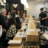 People gathered around a long wooden table with food items in a modern indoor setting.