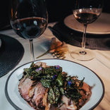 A plate of sliced cooked meat topped with herbs and flowers, set on a table with two glasses of red wine and place settings.