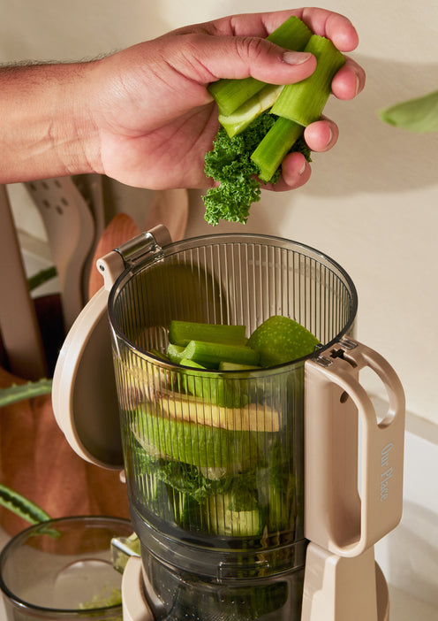 A hand places pieces of celery and parsley into a juicer filled with green vegetables on a kitchen counter.