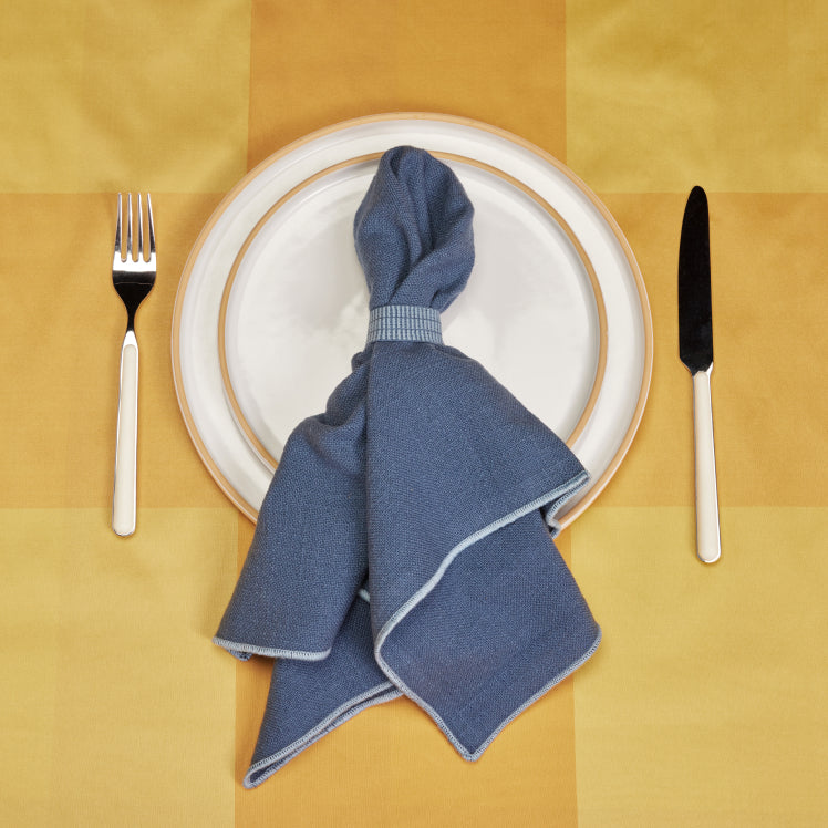 A blue Loop Napkin rests on top of beige dinnerware on a yellow tablecloth