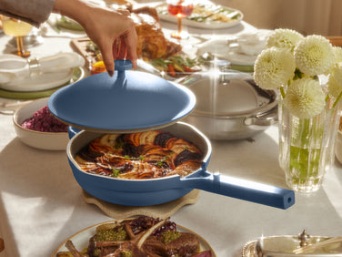 A person lifts the lid off a blue pan containing a baked dish, set on a dining table with various foods, flowers, and tableware.