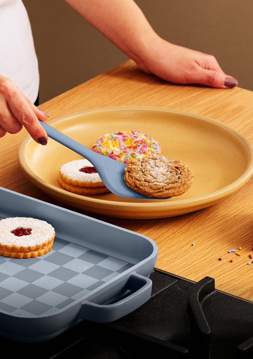 A hand uses a spatula to lift a cookie from a ceramic plate containing assorted cookies, next to a baking pan on a stovetop.