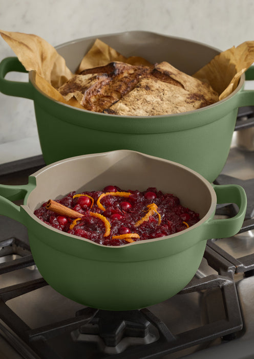 Two sage green cast iron Dutch ovens on a gas stovetop, with one filled with baked bread and the other with cranberry sauce, orange slices, and cinnamon.