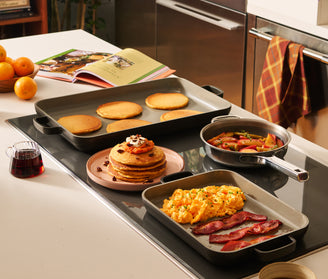Breakfast items including pancakes, eggs, and bacon on a stovetop with a cookbook in the background.