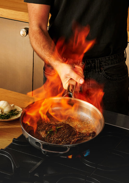 A person searing a steak in a stainless steel pan on a stovetop with high flames.