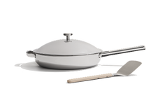 A stainless steel frying pan with a lid and a matching metal spatula with a light-colored handle, placed on a white background.