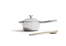 A white saucepan with a lid and long handle sits next to a beige wooden spoon on a plain white background.