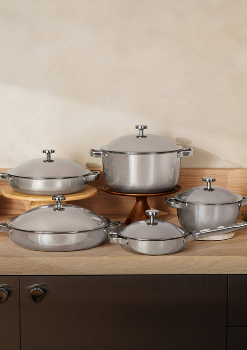 Five stainless steel pots and pans with lids are arranged on a wooden countertop against a beige wall.