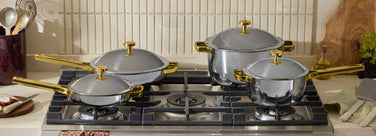 Four stainless steel pots and pans with gold handles and lids sit on a gas stove, surrounded by kitchen utensils and ingredients.