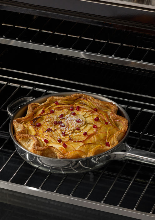 A round skillet with a golden-brown apple tart, topped with thin apple slices and scattered red berries, sits on the rack inside an oven.