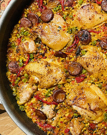 A close-up of a pan filled with paella, featuring golden chicken thighs, sliced chorizo, green peas, red bell peppers, and rice cooked with spices.