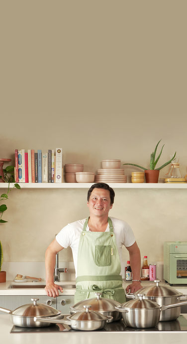 A person wearing a green apron stands in a modern kitchen, holding the lid of a stainless steel pan. Several stainless steel pots and pans are arranged on the counter in front of them.