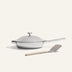 The Titanium Always Pan Pro, a white lidded frying pan with a long handle and innovative nonstick technology, sits next to a metal spatula with a beige handle on a light background.