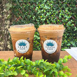 Two iced coffee drinks with frothy tops in clear plastic cups displaying the Agave Café logo sit on a wooden table. A green plant and lush leafy background add a fresh, vibrant atmosphere.