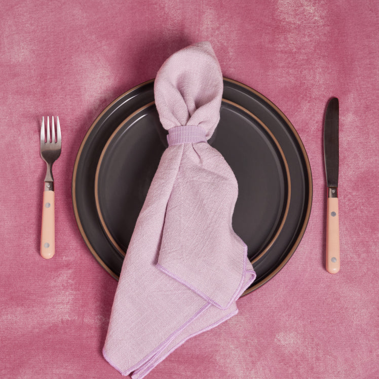 A purple Loop Napkin rests on top of black dinnerware on a pink tablecloth