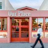 A person walks past the storefront of Our Place, a shop with a salmon-pink exterior, large windows displaying cookware, and bold white lettering above the entrance.