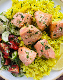 A plate with yellow rice, herb-seasoned salmon chunks, cucumber-tomato salad, and a lemon wedge.