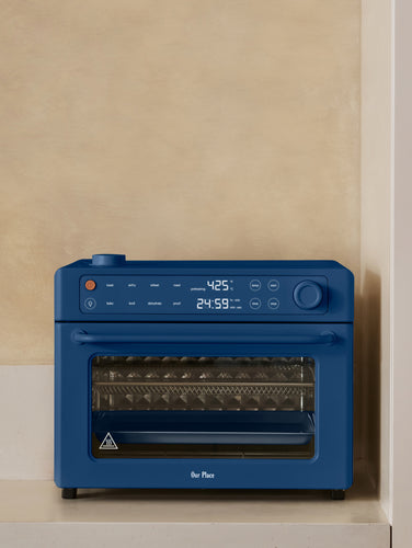 Blue toaster oven on a kitchen counter with a vase, bottles, and a plant in the background.