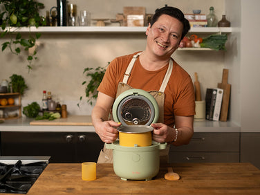 A person in an orange shirt and apron stands in a kitchen, smiling while placing a yellow container into a green rice cooker on the counter.
