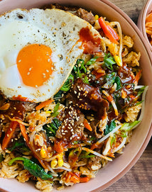 A bowl of bibimbap topped with a sunny-side-up egg, vegetables, beef, rice, and sesame seeds, drizzled with sauce.