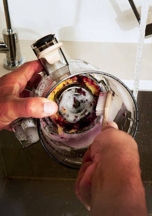 A person scrubs the inside of a soapy food processor bowl under running water at a kitchen sink.